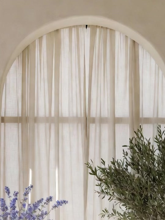 Sheer linen curtains glowing in a tall arched window, with olive leaves at the edge